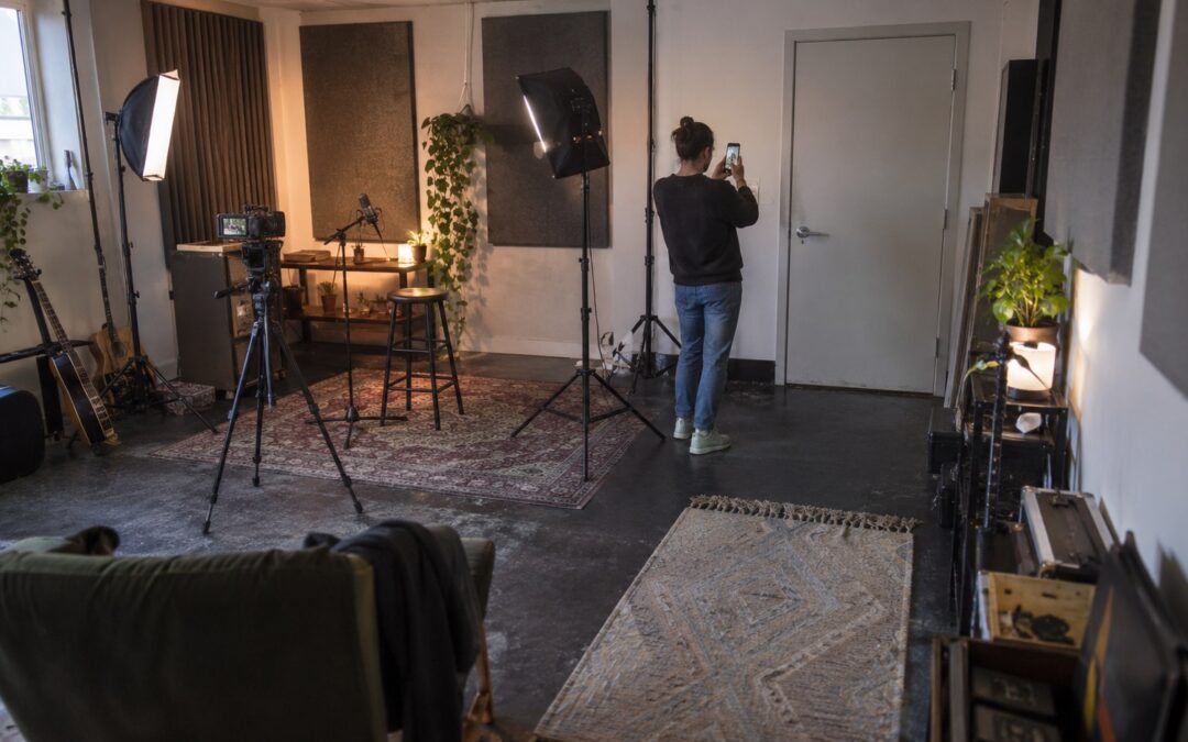 A performer photographs a closed door in a converted rehearsal studio with a camera rig, microphones, acoustic panels, and lighting stands.
