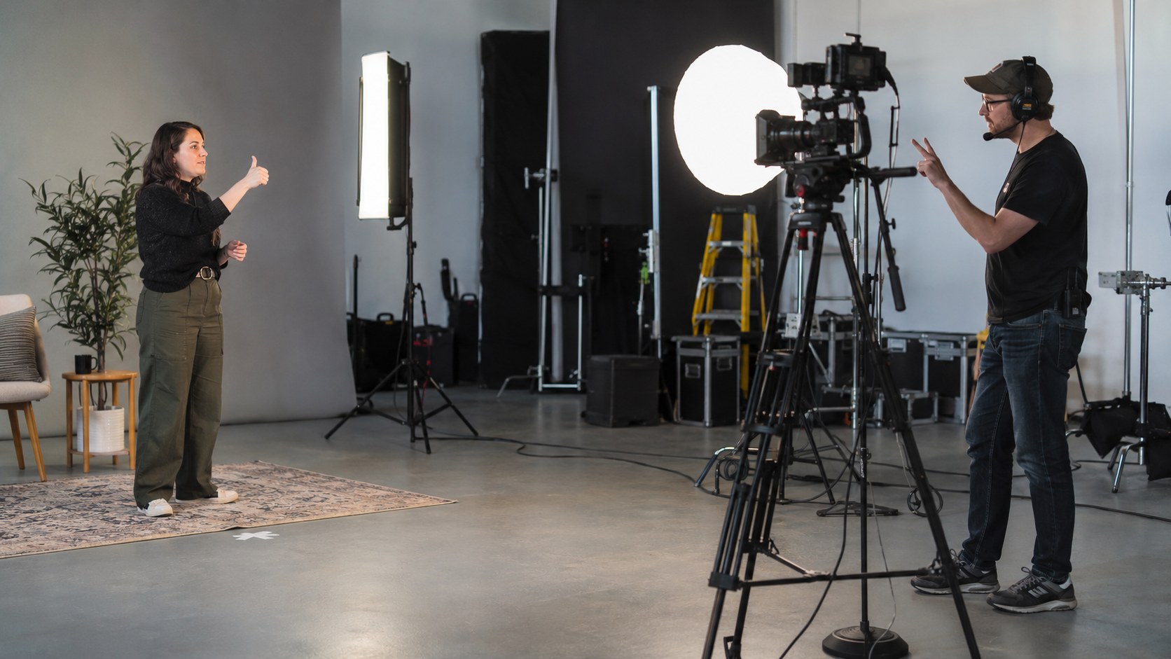 A performer and moderator exchange hand signals across a neutral video studio beside a lit camera and production.