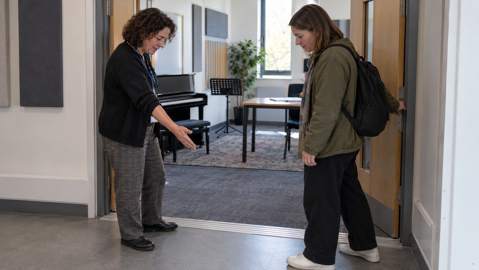 A tutor holds open a teaching studio door while a support coordinator gestures toward the threshold during an access check.